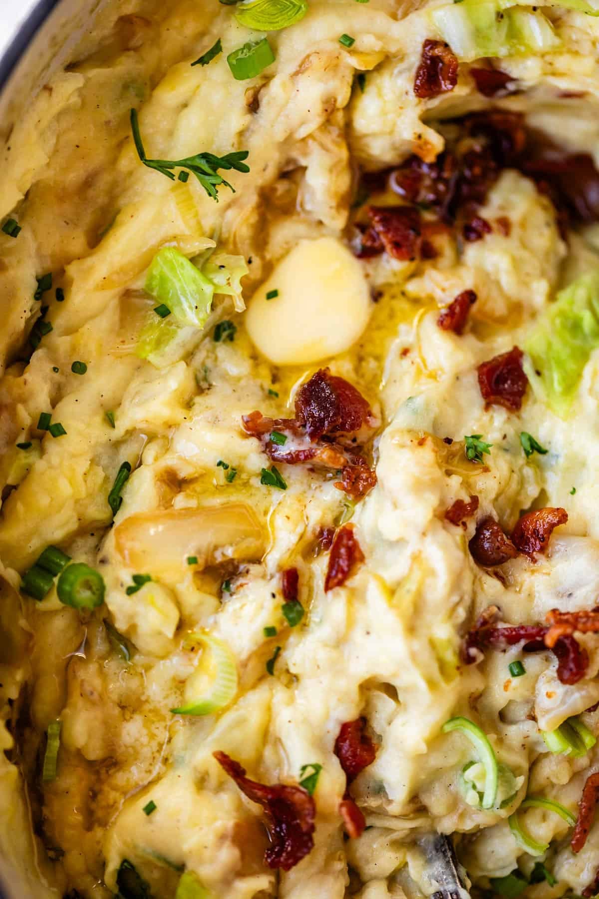 Close-up of creamy mashed potatoes topped with melted butter, chopped green onions, crispy bacon bits, herbs, and visible roasted garlic cloves.