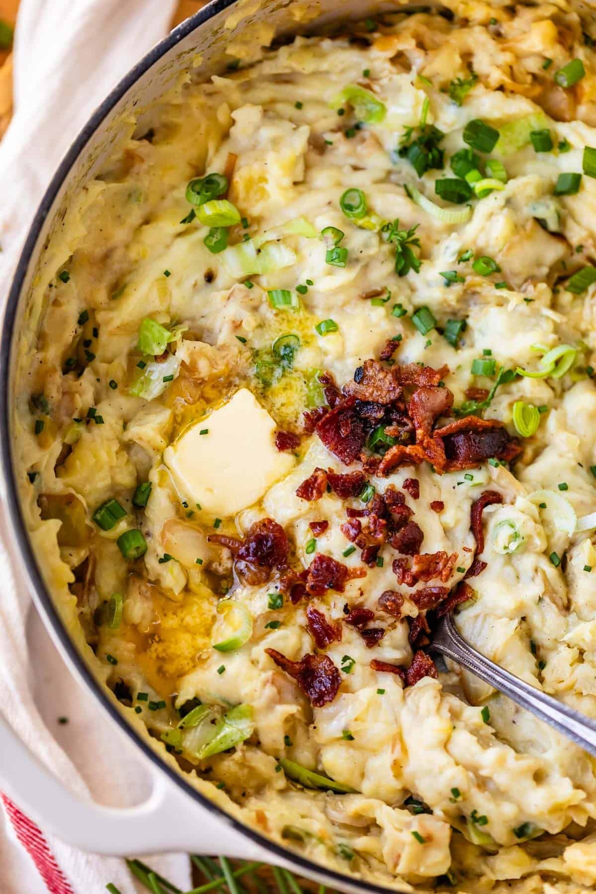A pot of creamy mashed potatoes topped with melted butter, crispy bacon bits, and chopped green onions, with a spoon partially dipped in the mixture.
