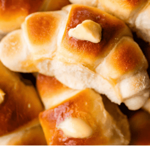 A close-up of golden brown crescent rolls with pats of melting butter on top. The rolls look soft and fluffy, and the text below reads Soft & Pillowy Crescent Rolls - The Food Charlatan.