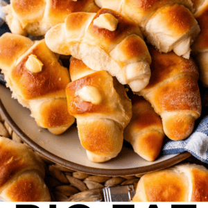 A plate piled with golden-brown crescent rolls topped with melted butter, surrounded by more rolls on a woven tray. The text at the bottom reads, Big Fat Crescent Rolls - The Food Charlatan.