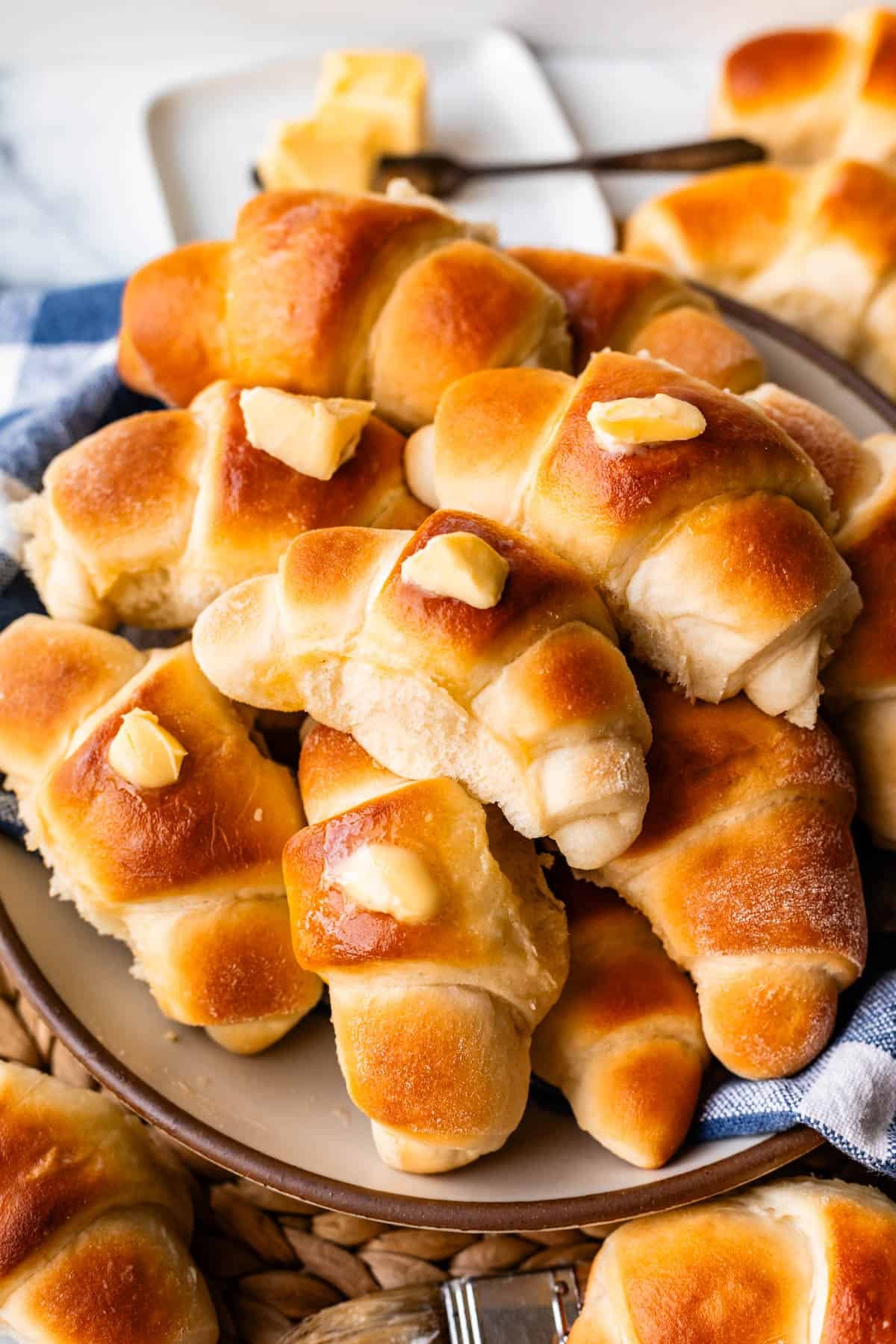 A plate stacked with golden, freshly baked crescent rolls, each topped with a pat of melting butter, with more butter on a dish in the background.