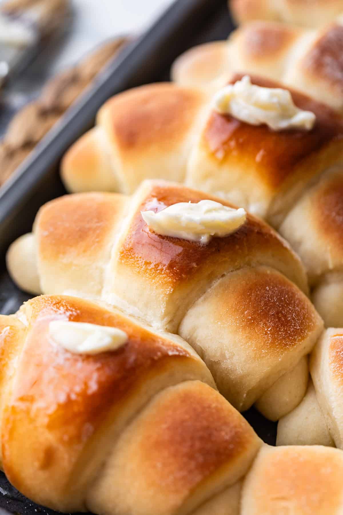 Freshly baked golden-brown crescent rolls topped with melting pats of butter, arranged closely together on a dark baking tray.