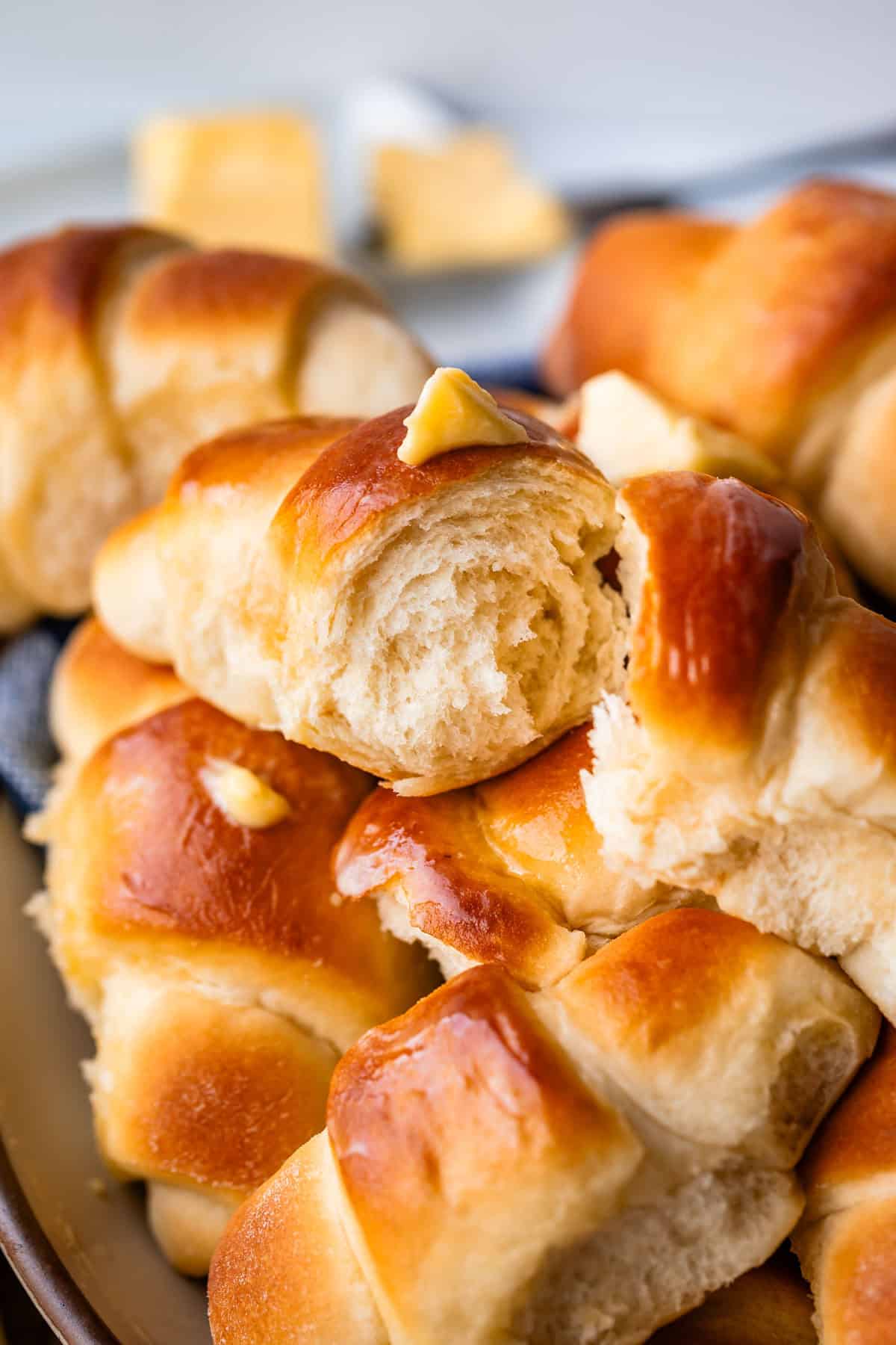 A close-up of freshly baked golden-brown crescent rolls, with a pat of butter melting on top and more butter blurred in the background. The rolls look soft, fluffy, and slightly shiny.