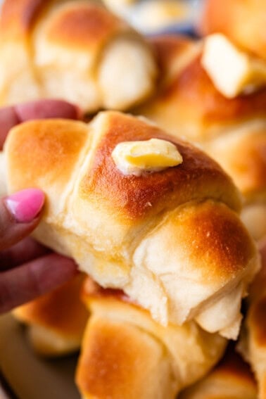 A close-up of a hand holding a golden brown, fluffy dinner roll topped with a pat of melting butter, with more rolls stacked in the background.