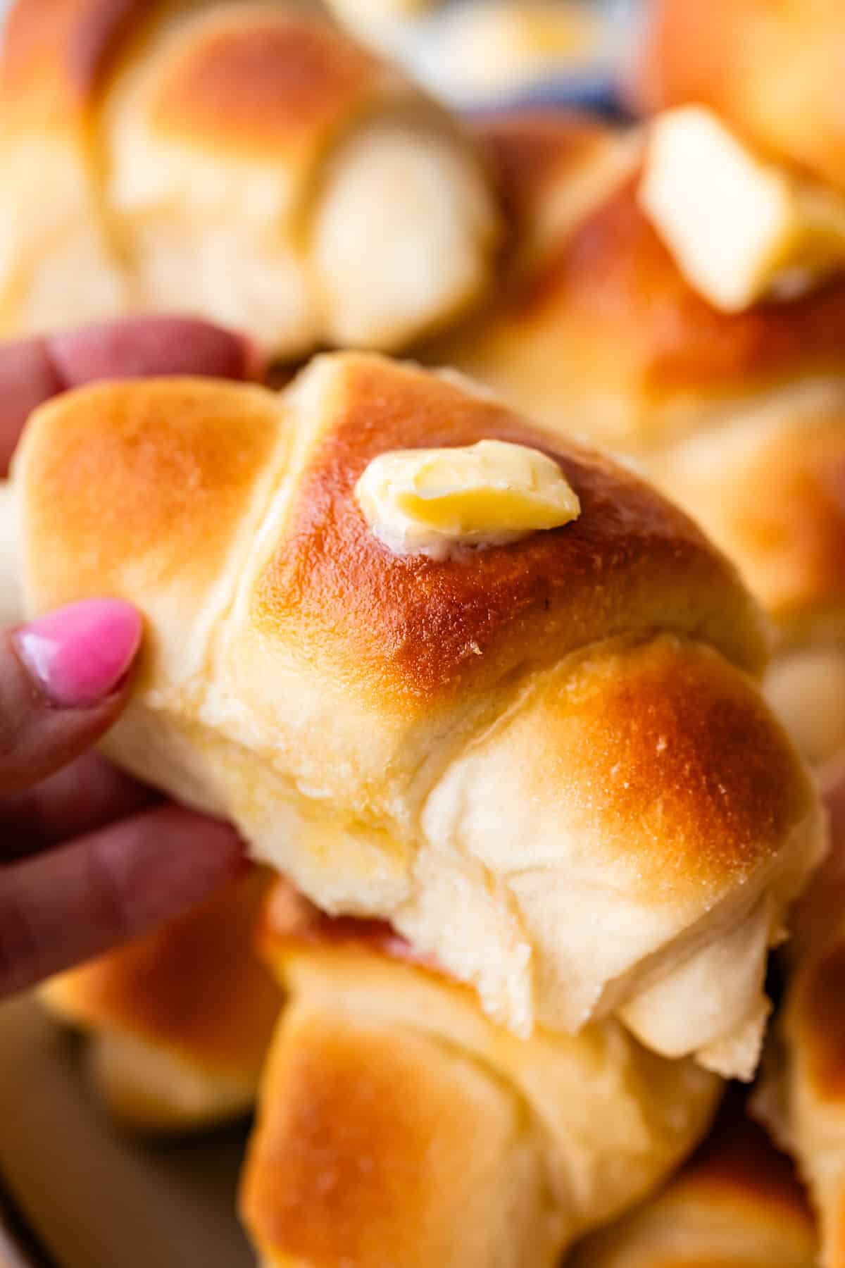 A close-up of a hand holding a golden brown, fluffy dinner roll topped with a pat of melting butter, with more rolls stacked in the background.