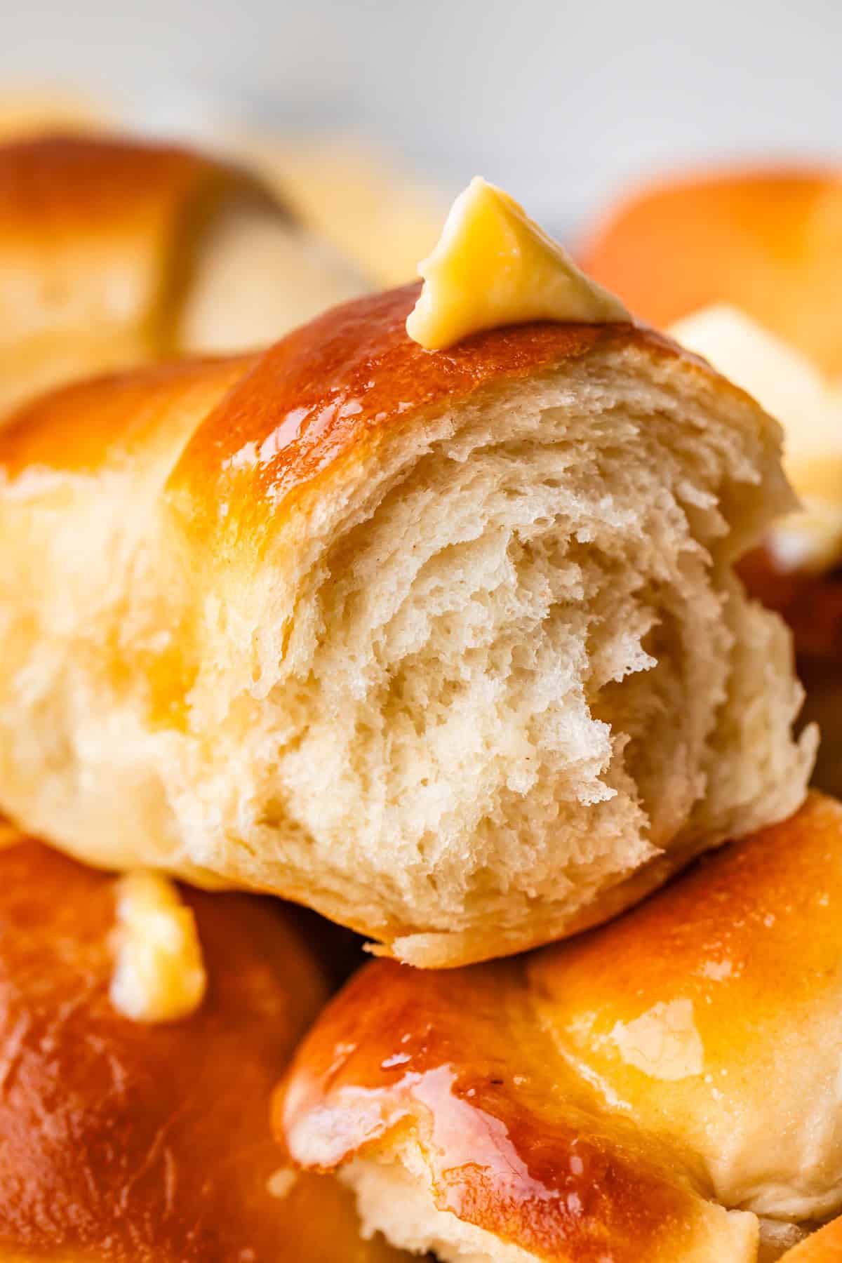 A close-up of a soft, fluffy dinner roll with a shiny, golden-brown top and a pat of butter melting on it. Other rolls are visible in the background.