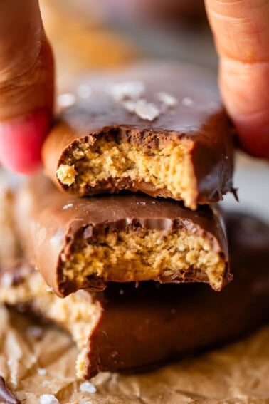 A close-up of a hand holding a chocolate-coated peanut butter egg with a bite taken out, stacked on top of another partially eaten cookie, both showing a crumbly peanut butter filling.