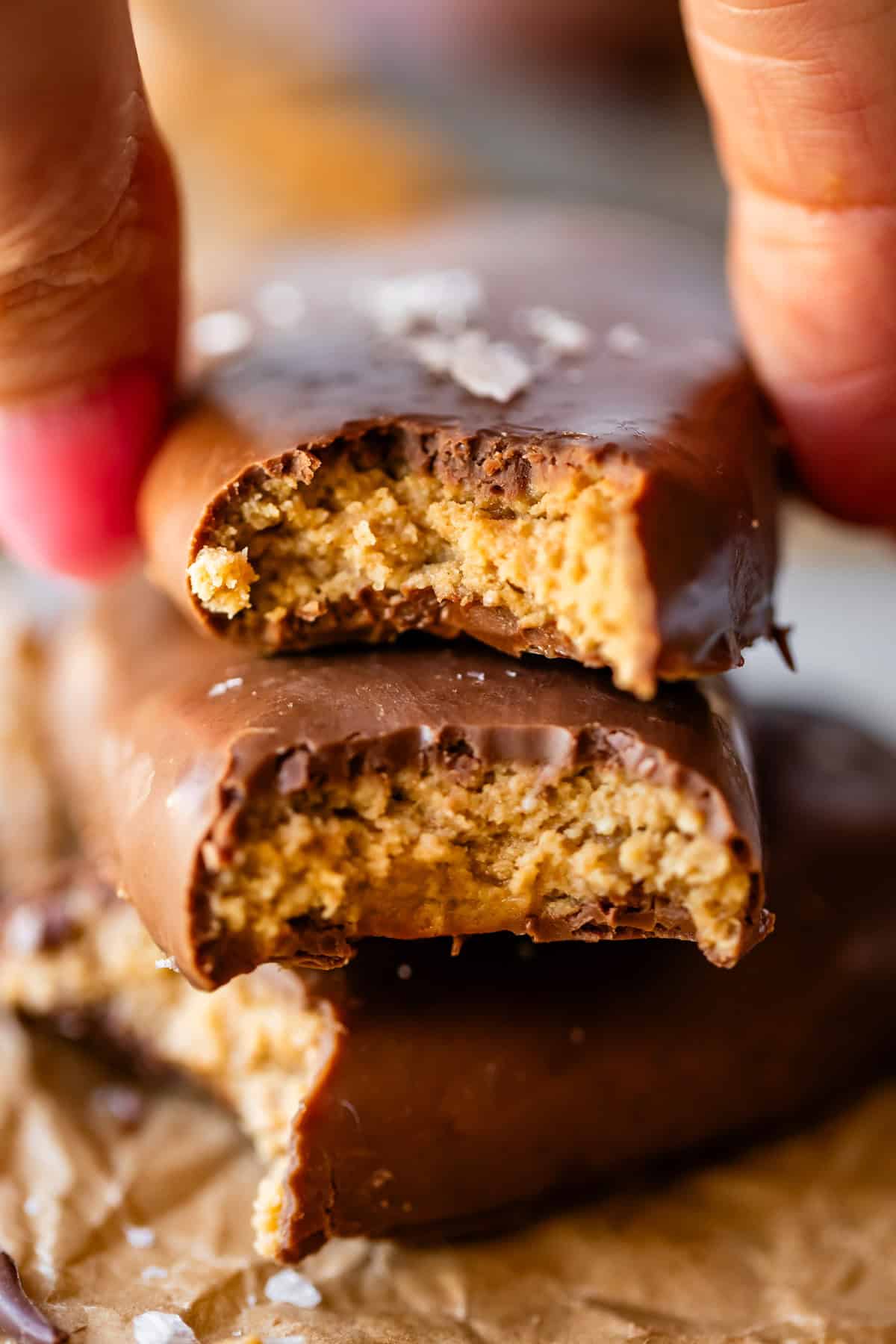 A close-up of a hand holding a chocolate-coated peanut butter egg with a bite taken out, stacked on top of another partially eaten cookie, both showing a crumbly peanut butter filling.
