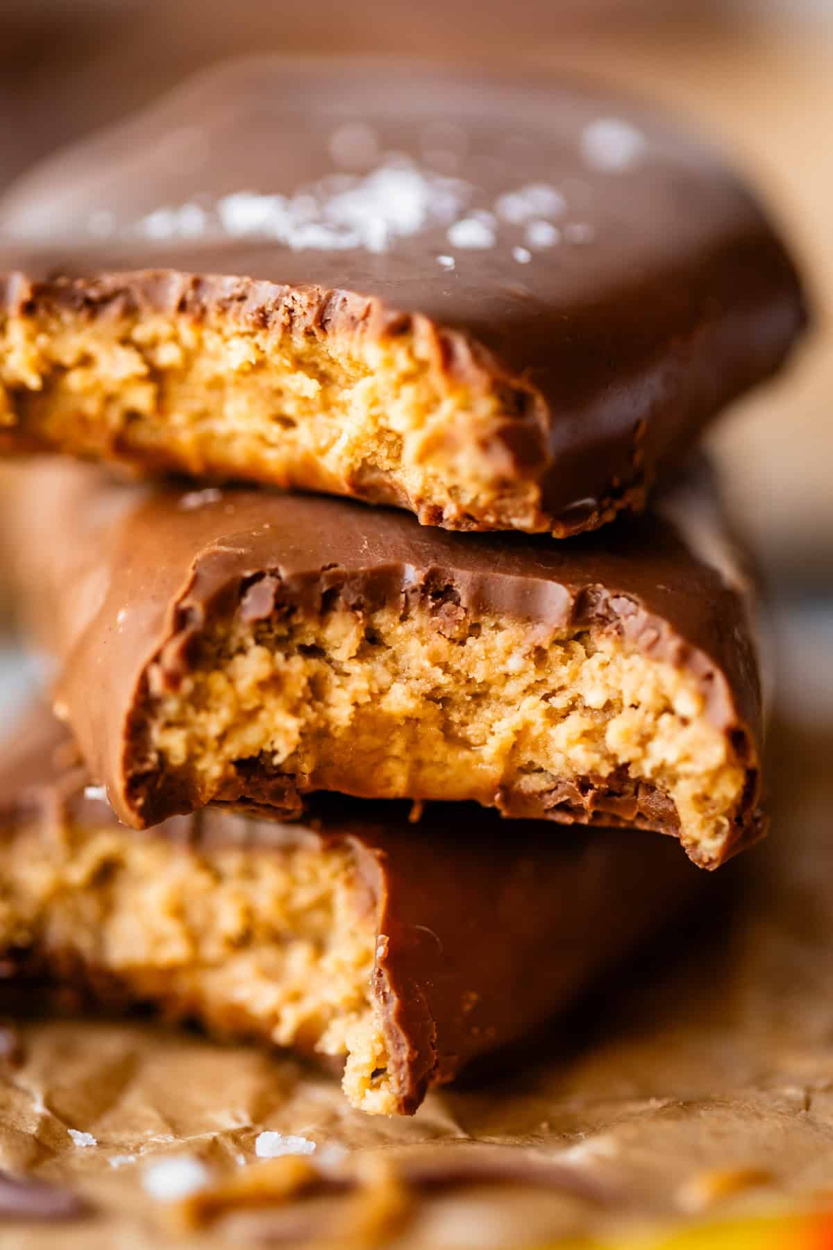 Two chocolate-covered peanut butter eggs are stacked, both with large bites taken out, revealing a creamy peanut butter filling inside. The bars rest on a crumpled piece of brown parchment paper.