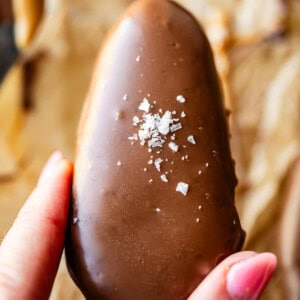 A hand with pink nail polish holds a chocolate-covered peanut butter egg topped with a sprinkle of coarse sea salt, against a blurred background of parchment paper and other treats.