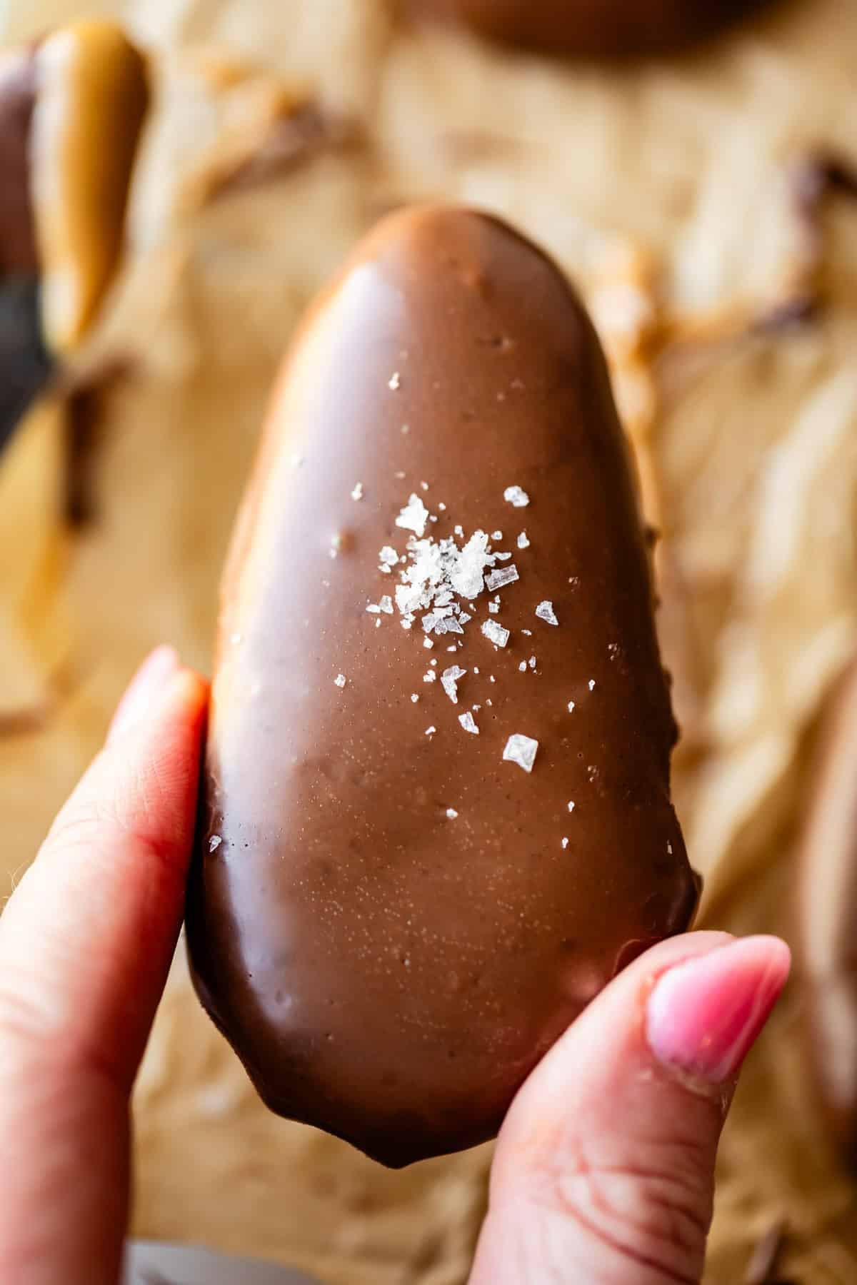 A hand with pink nail polish holds a chocolate-covered peanut butter egg topped with a sprinkle of coarse sea salt, against a blurred background of parchment paper and other treats.