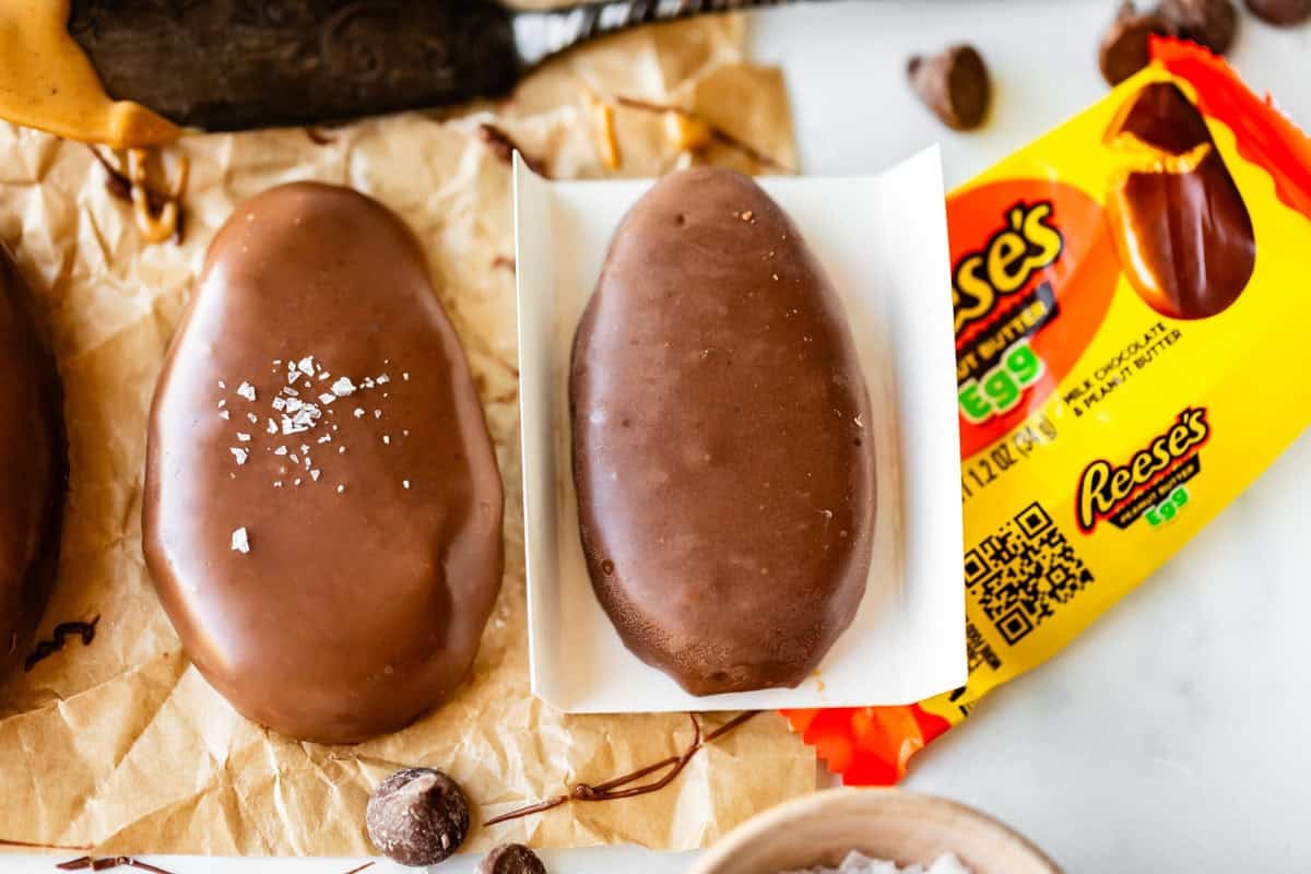 A close-up of two chocolate-covered Reese’s eggs, one sprinkled with sea salt, next to an open yellow Reese’s Egg wrapper on crumpled parchment paper. Chocolate chips and chocolate smudges are nearby.