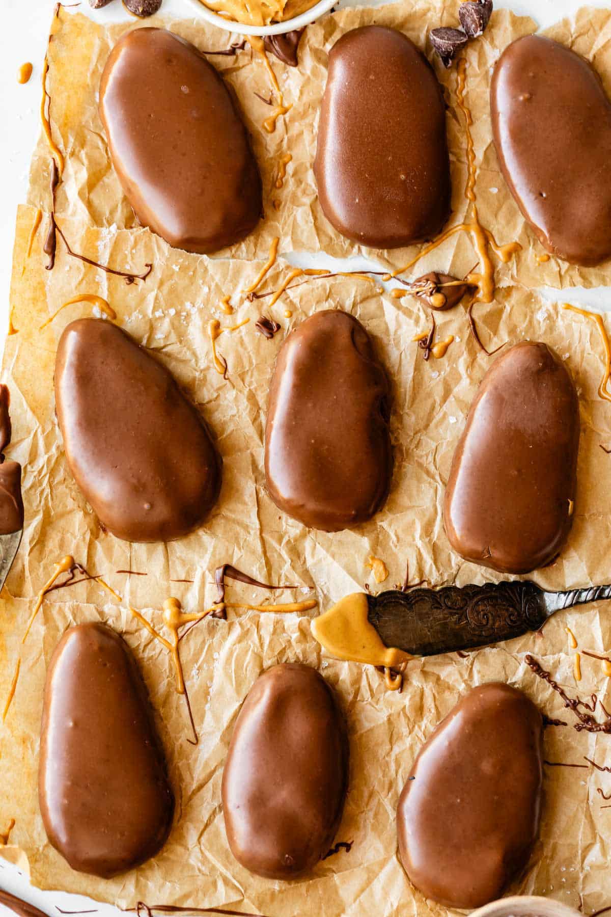 Nine oval-shaped chocolate-covered peanut butter eggs are arranged on crumpled parchment paper, with some chocolate and peanut butter smudges. A small knife with peanut butter rests nearby.
