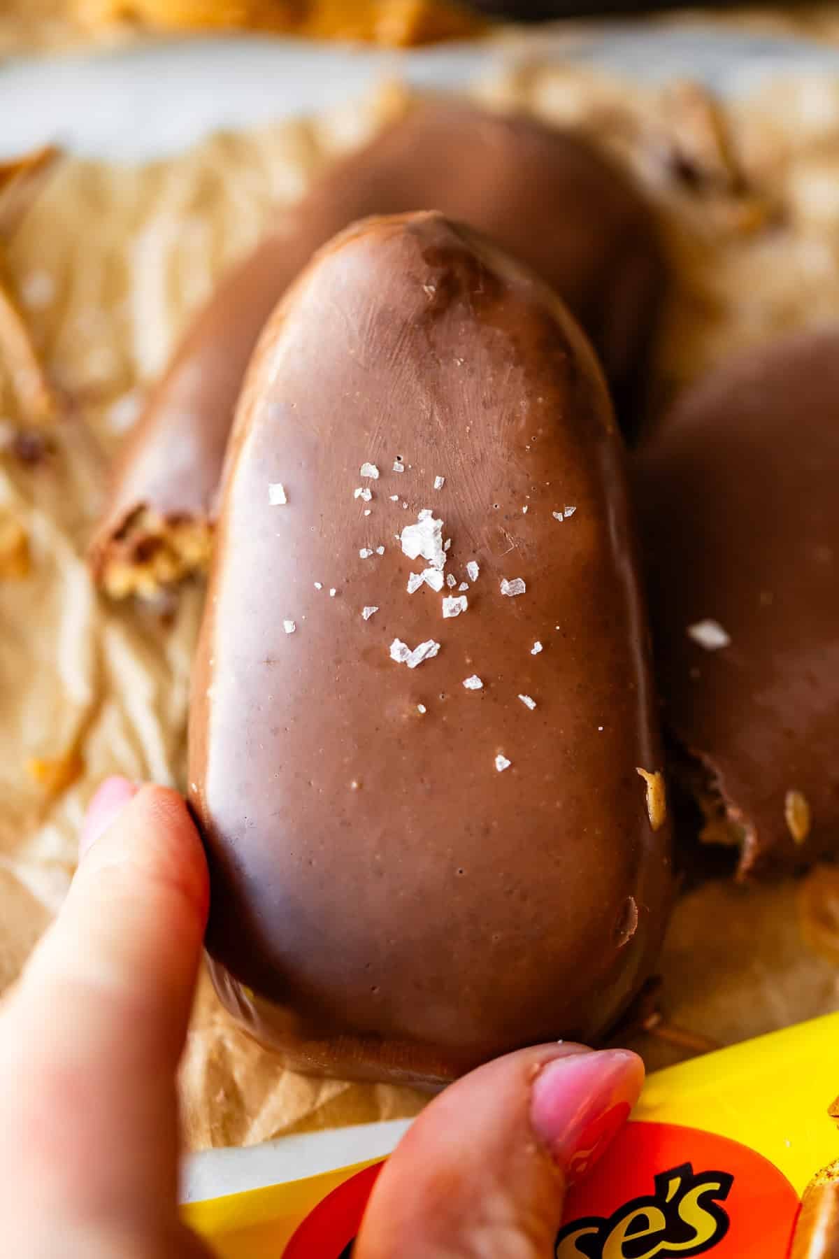 A hand holds a chocolate-covered peanut butter egg topped with flaky sea salt, resting on brown parchment paper with a partially visible Reese’s wrapper below.