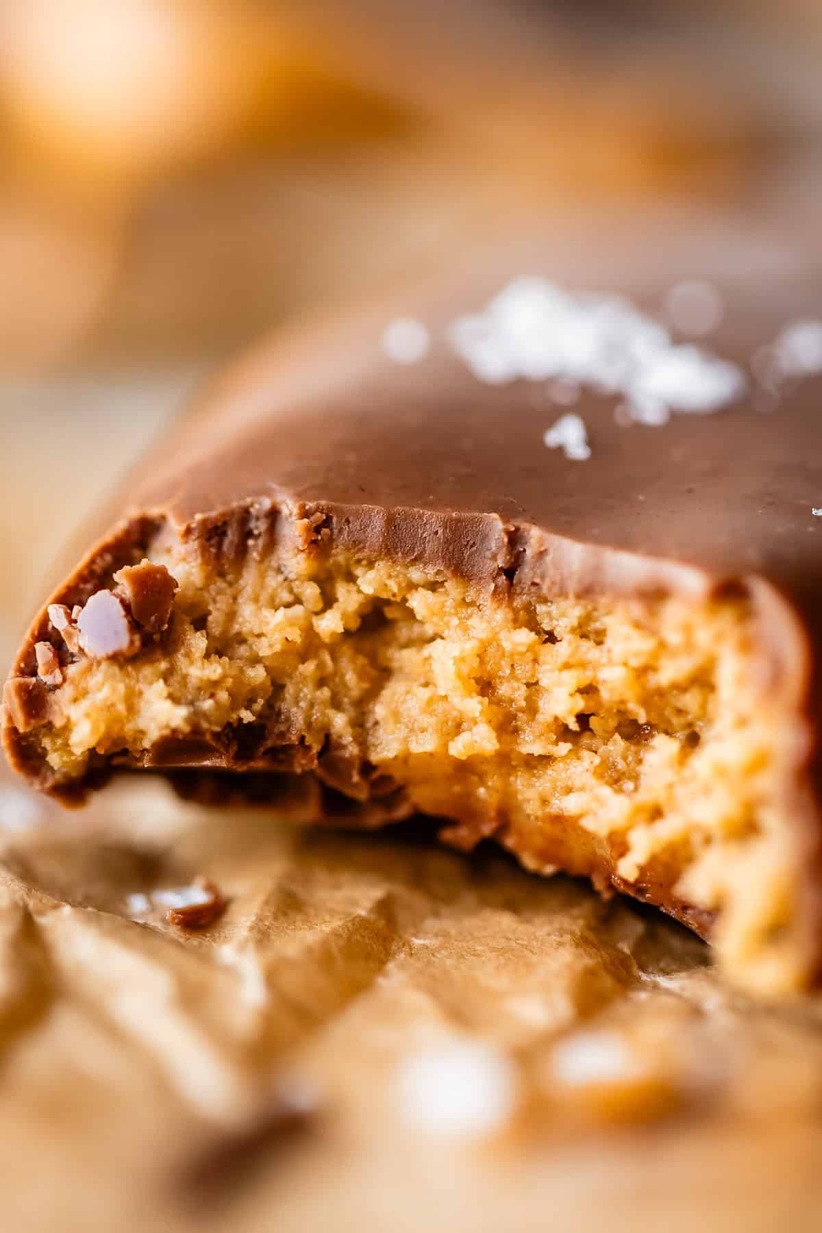 A close-up of a chocolate-covered peanut butter egg with a bite taken out, revealing a creamy peanut butter filling. The bar rests on crinkled brown parchment paper with a sprinkle of sea salt on top.