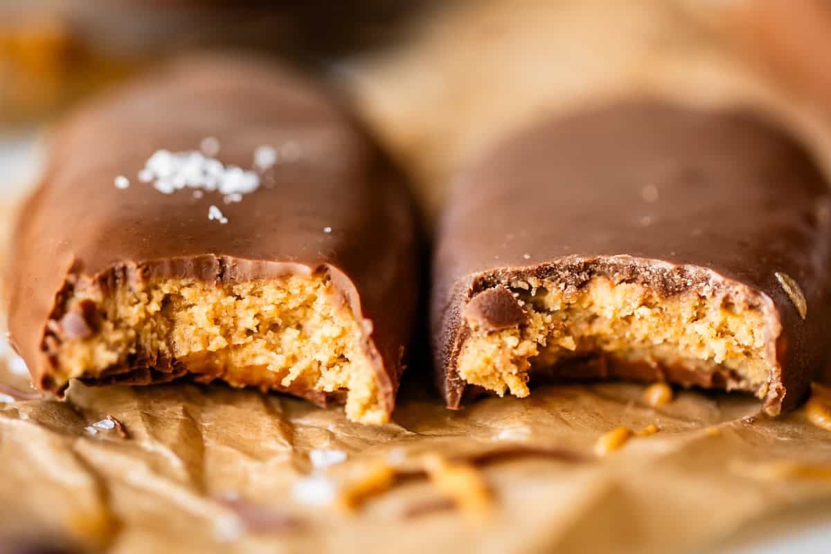 Two chocolate-coated peanut butter bars, each with a bite taken out, revealing a creamy peanut butter filling. They rest on crumpled brown parchment paper, with a sprinkle of salt on one chocolate coating.