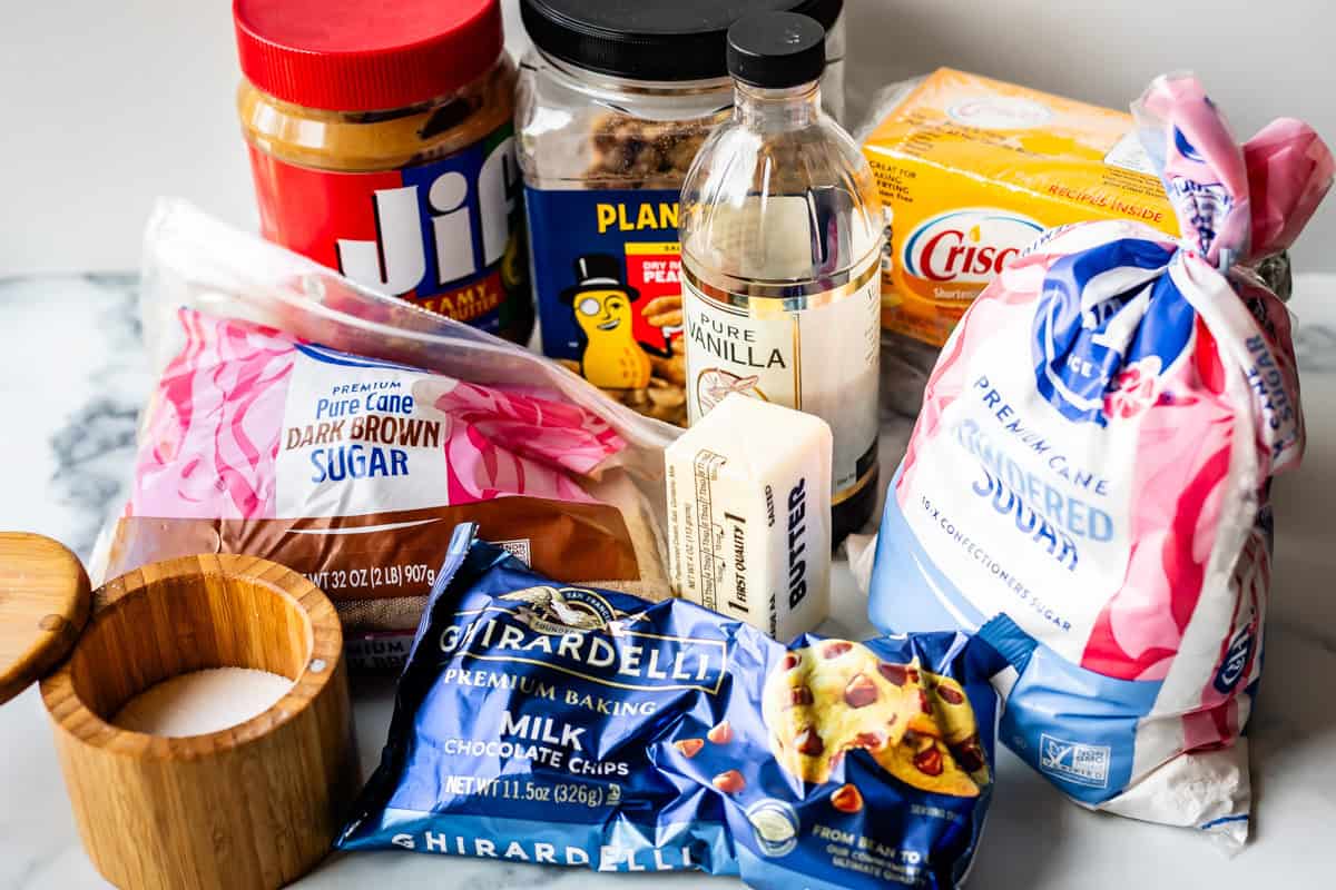 A variety of baking ingredients on a counter, including peanut butter, brown sugar, powdered sugar, chocolate chips, vanilla extract, butter, shortening, white sugar in a wooden container, and a bag of flour.