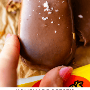 A hand holding a chocolate-covered peanut butter egg topped with sea salt, resting on parchment paper. Text reads: Homemade Reese’s Peanut Butter Eggs - The Food Charlatan.