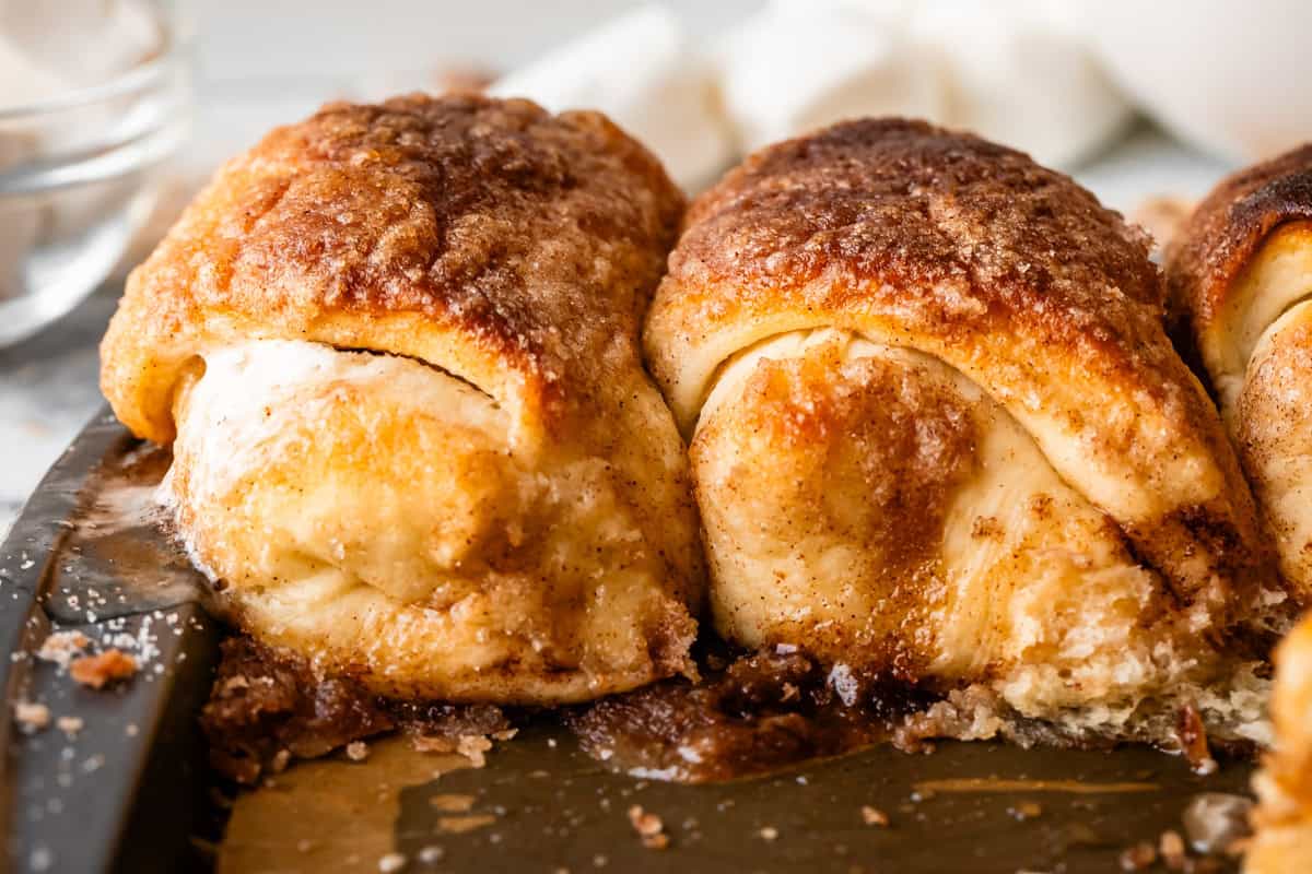 Close-up of golden-brown crusted resurrection rolls with gooey marshmallow filling, sitting on a baking tray with melted sugar and spices around them.