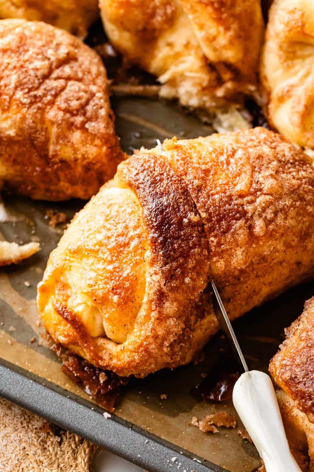 A close-up of a golden-brown, cinnamon-sugar-coated empty tomb roll being sliced with a knife on a baking sheet, surrounded by other similar pastries.
