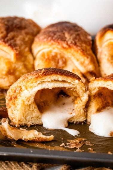 Close-up of golden-brown resurrection crescent rolls, with one roll cut open to reveal a gooey, melted marshmallow filling oozing out onto the baking sheet.