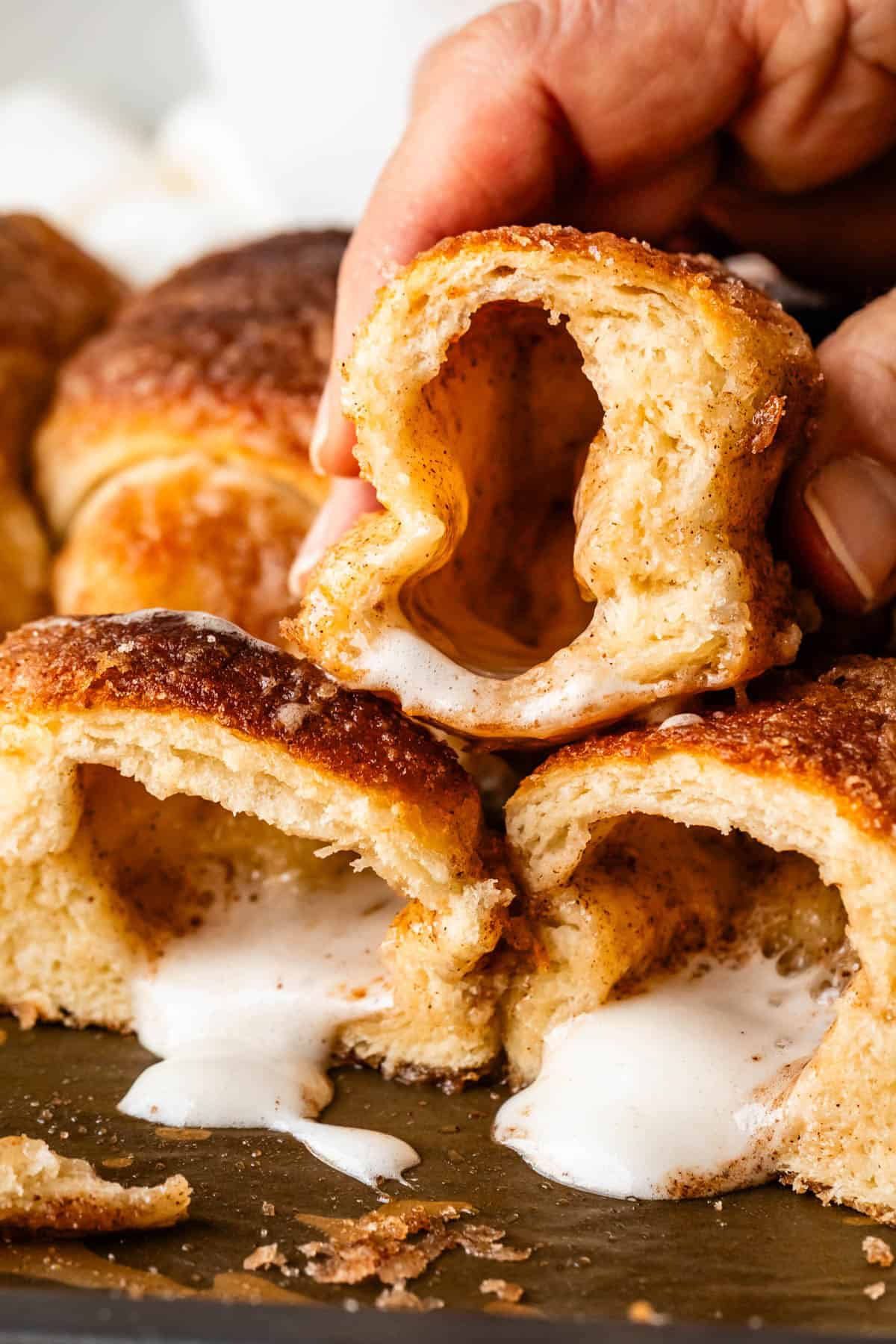 A hand holds a cinnamon sugar pastry, revealing a gooey, melted marshmallow center. More pastries with oozing marshmallow filling are stacked below on a tray.