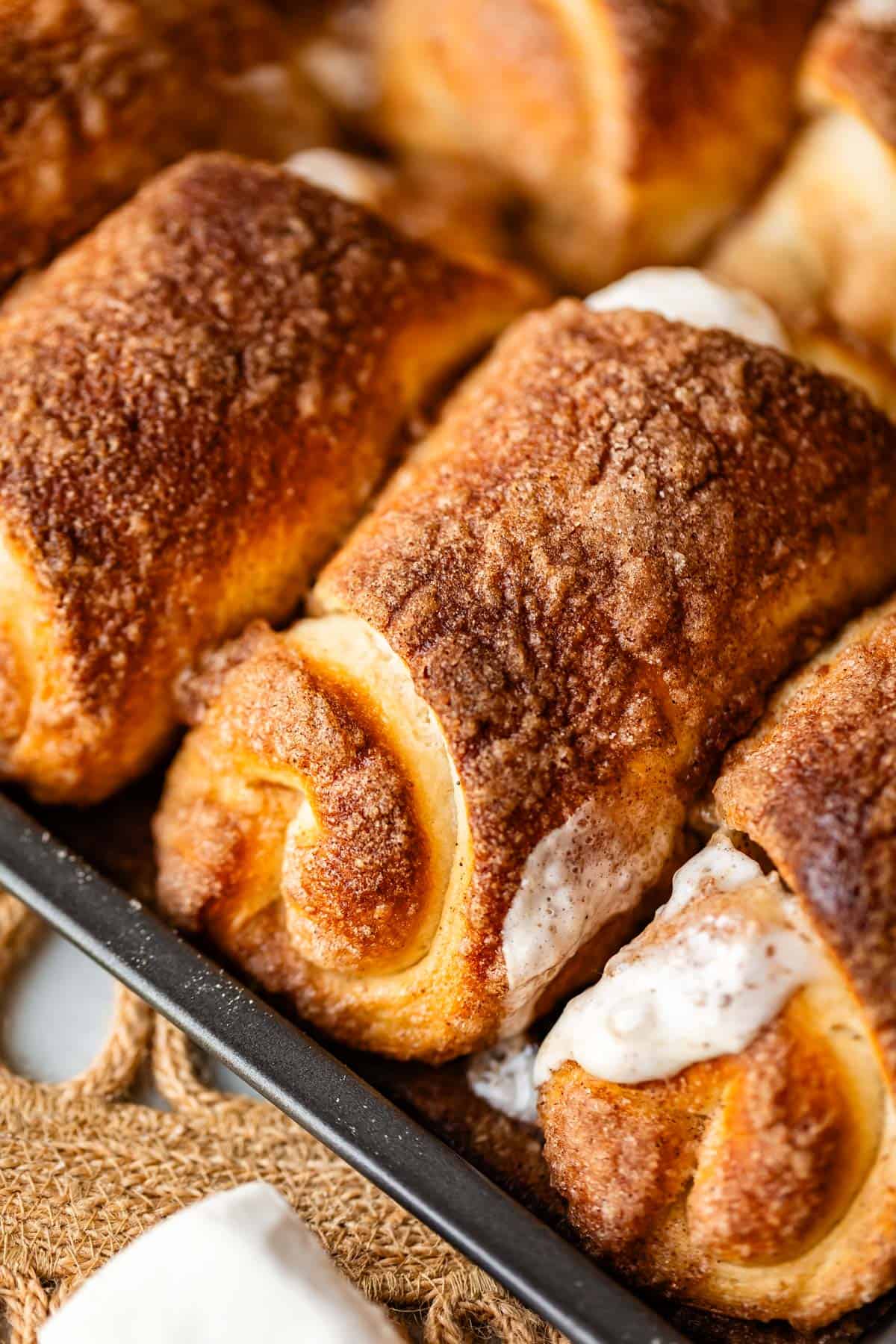 Close-up of golden, cinnamon-sugar coated resurrection rolls in a baking tray, with some creamy white icing melting between the rolls.
