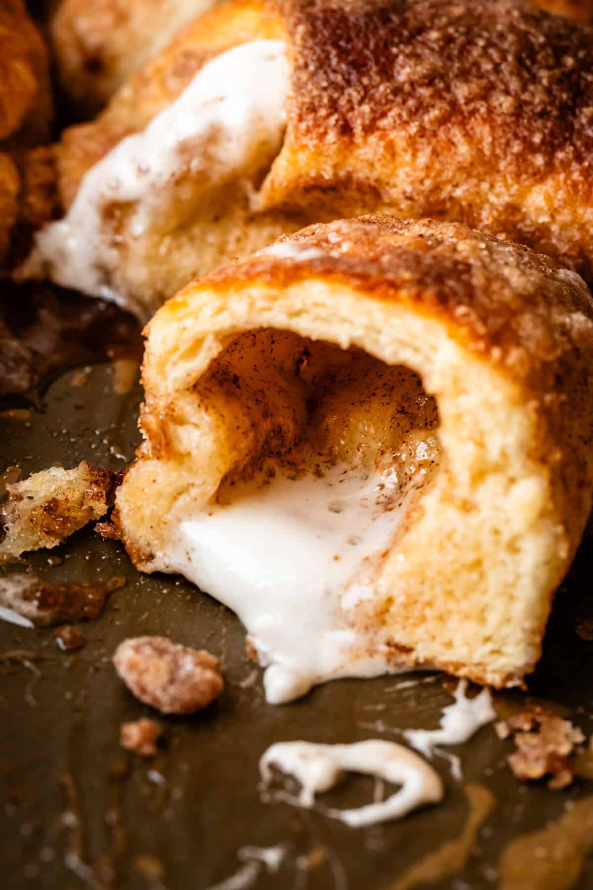 A close-up of a baked resurrection roll with a gooey, melted marshmallow filling oozing out from the center, surrounded by crumbs and a golden, cinnamon-sugar crust.