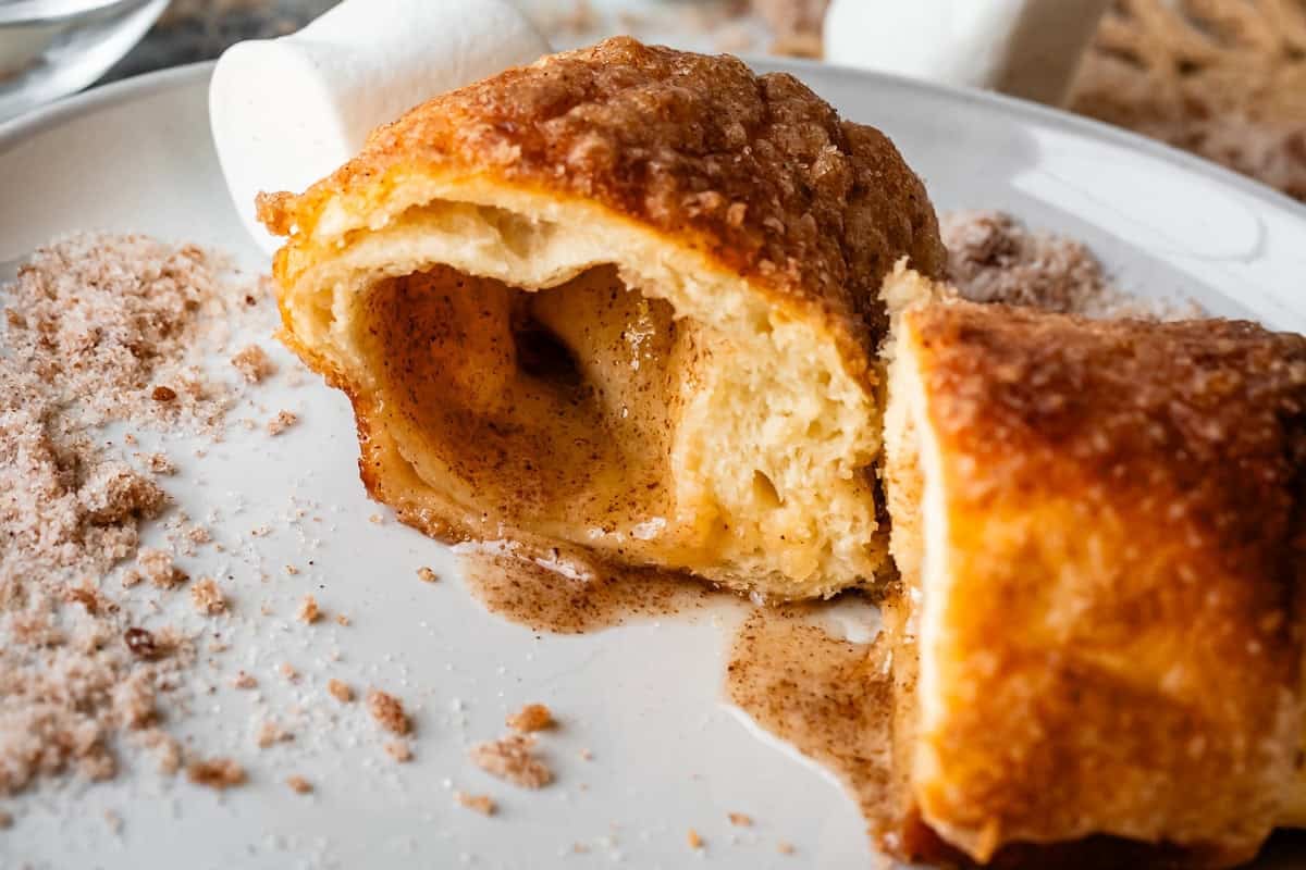A close-up of a resurrection roll with a golden, flaky crust, split open to reveal a gooey cinnamon sugar filling, with sugar and cinnamon sprinkled on the white plate beside it.