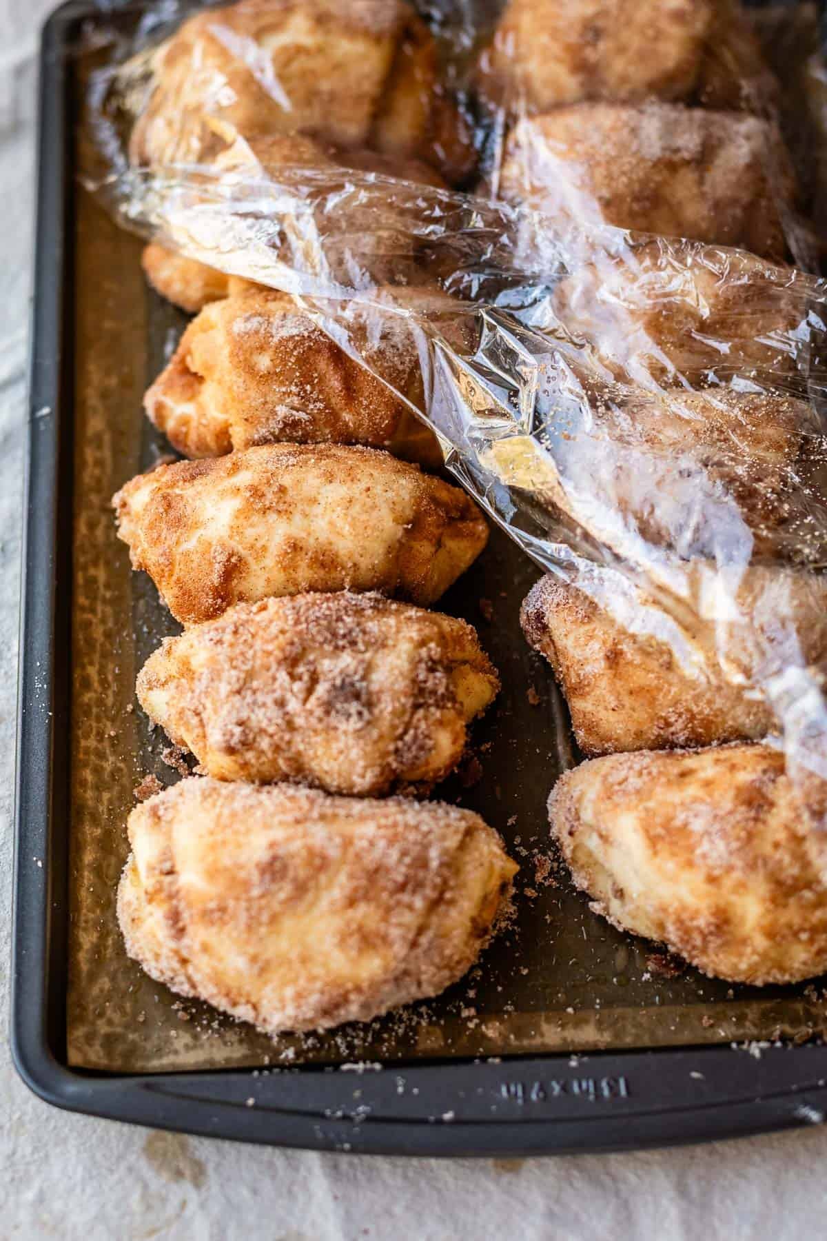 A baking sheet filled with unbaked, crescent-shaped dough rolls coated in cinnamon sugar, partially covered with plastic wrap, ready to be baked.