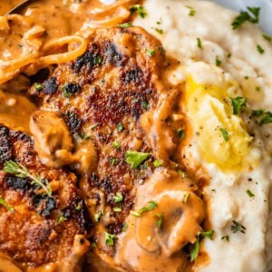 A close-up of creamy mashed potatoes with a pat of butter, served with pan-seared pork chops topped with a rich brown mushroom gravy, garnished with chopped parsley.