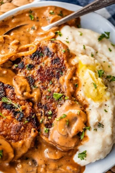 A close-up of creamy mashed potatoes with a pat of butter, served with pan-seared pork chops topped with a rich brown mushroom gravy, garnished with chopped parsley.