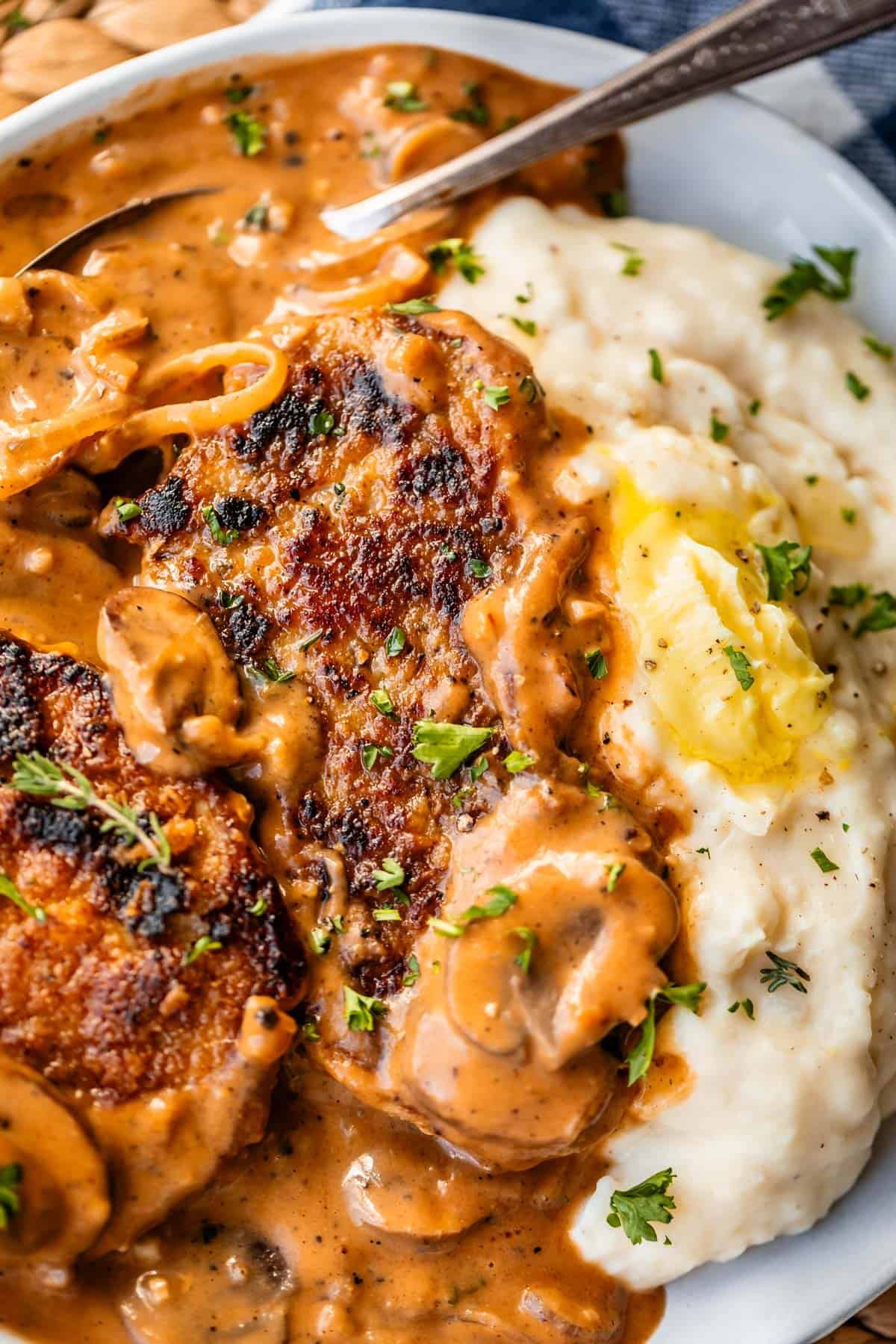 A close-up of creamy mashed potatoes with a pat of butter, served with pan-seared pork chops topped with a rich brown mushroom gravy, garnished with chopped parsley.