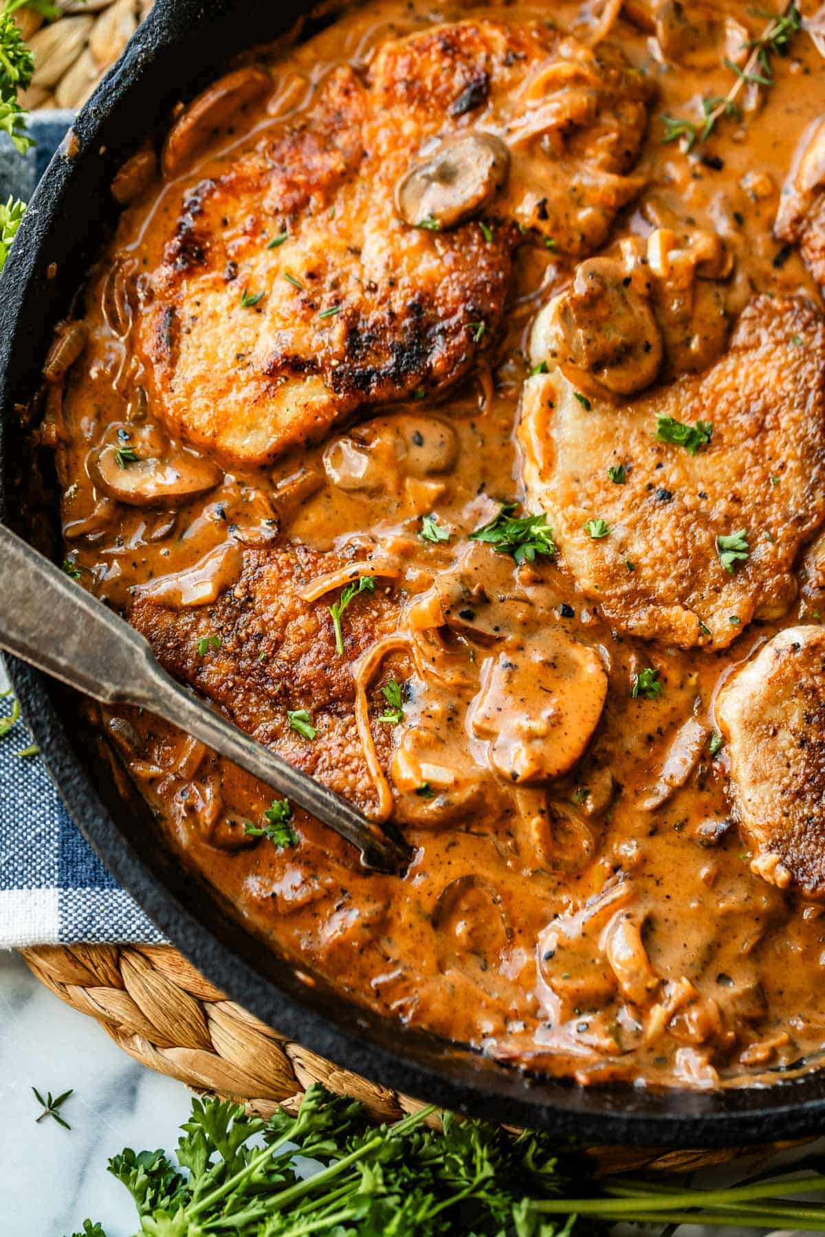 A skillet filled with golden-brown pork chops in a creamy mushroom sauce, garnished with chopped parsley. A spoon rests in the skillet, and fresh herbs are visible nearby.