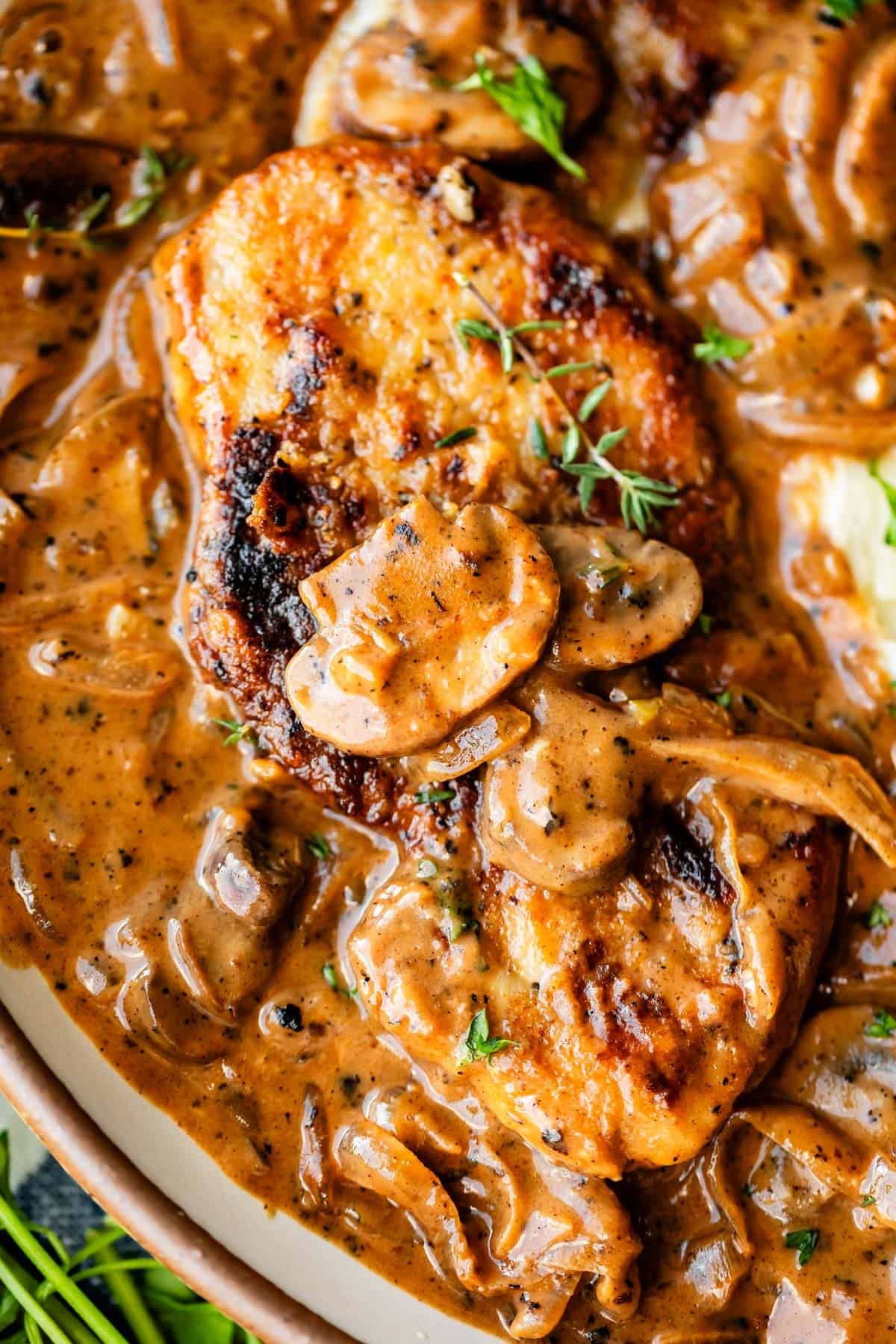  A close-up of a juicy, golden-brown grilled chicken breast topped with sliced mushrooms and herbs, covered in a creamy brown sauce with onions and garnished with a sprig of fresh thyme.