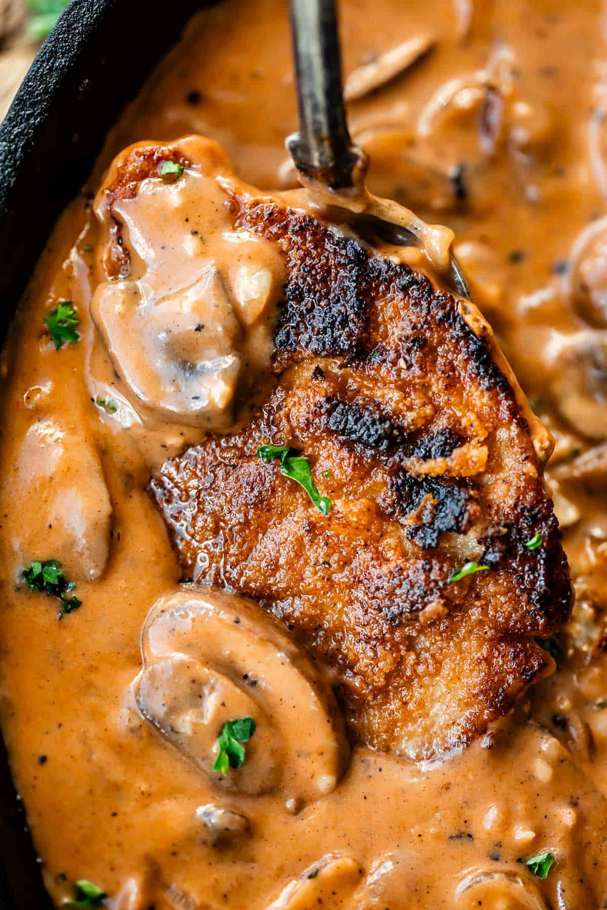 A golden-brown, crispy pork chop topped with creamy mushroom sauce and garnished with parsley, served in a skillet.