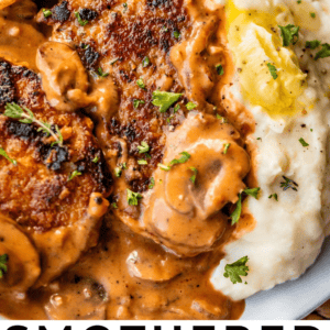 A close-up of smothered pork chops topped with creamy mushroom gravy, served alongside mashed potatoes with herbs, on a white plate. Text at the bottom reads, Smothered Pork Chops - The Food Charlatan.