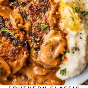 A close-up of smothered pork chops topped with creamy brown gravy and fresh herbs, served alongside creamy mashed potatoes with melted butter. Text reads: Southern Classic Smothered Pork Chops – The Food Charlatan.