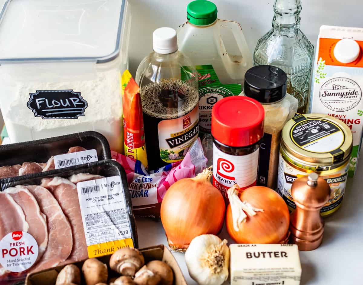 A variety of cooking ingredients on a counter, including pork chops, onions, garlic, mushrooms, flour, sugar, soy sauce, vinegar, broth, heavy cream, butter, pepper grinder, and seasonings.