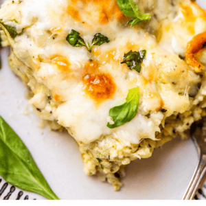 A close-up of a slice of white chicken lasagna topped with melted cheese, fresh basil, and herbs on a plate with a spoon. The text below reads White Chicken Lasagna - The Food Charlatan.