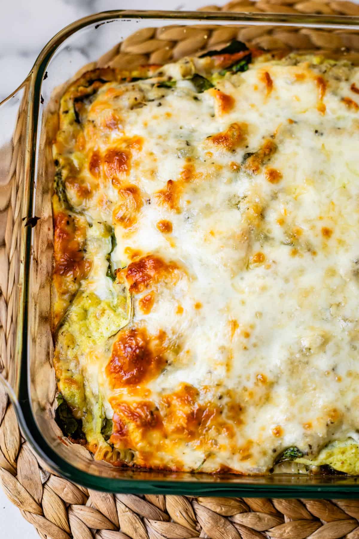 A close-up of a baked zucchini lasagna in a glass dish, topped with melted, golden-brown cheese, resting on a woven placemat.