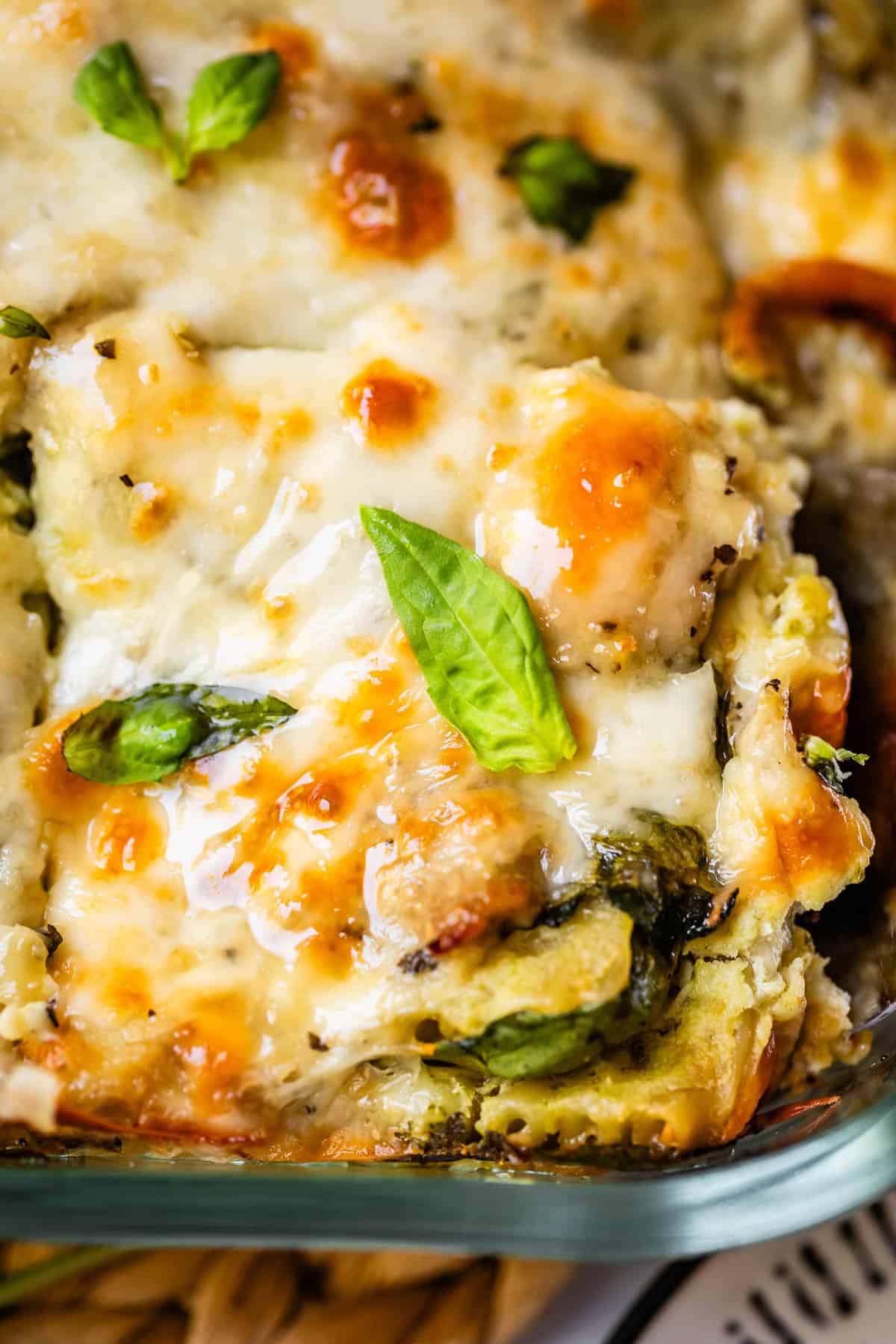 Close-up of a cheesy, baked vegetable lasagna topped with melted golden cheese and fresh basil leaves, showing layers of pasta and greens in a glass baking dish.