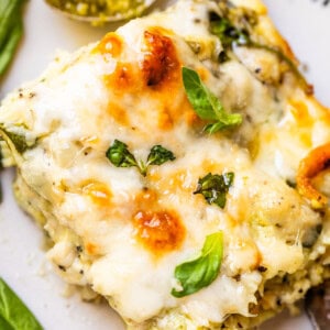 A close-up of a slice of cheesy lasagna topped with melted cheese and fresh basil leaves on a white plate, with a spoonful of green pesto sauce in the background.