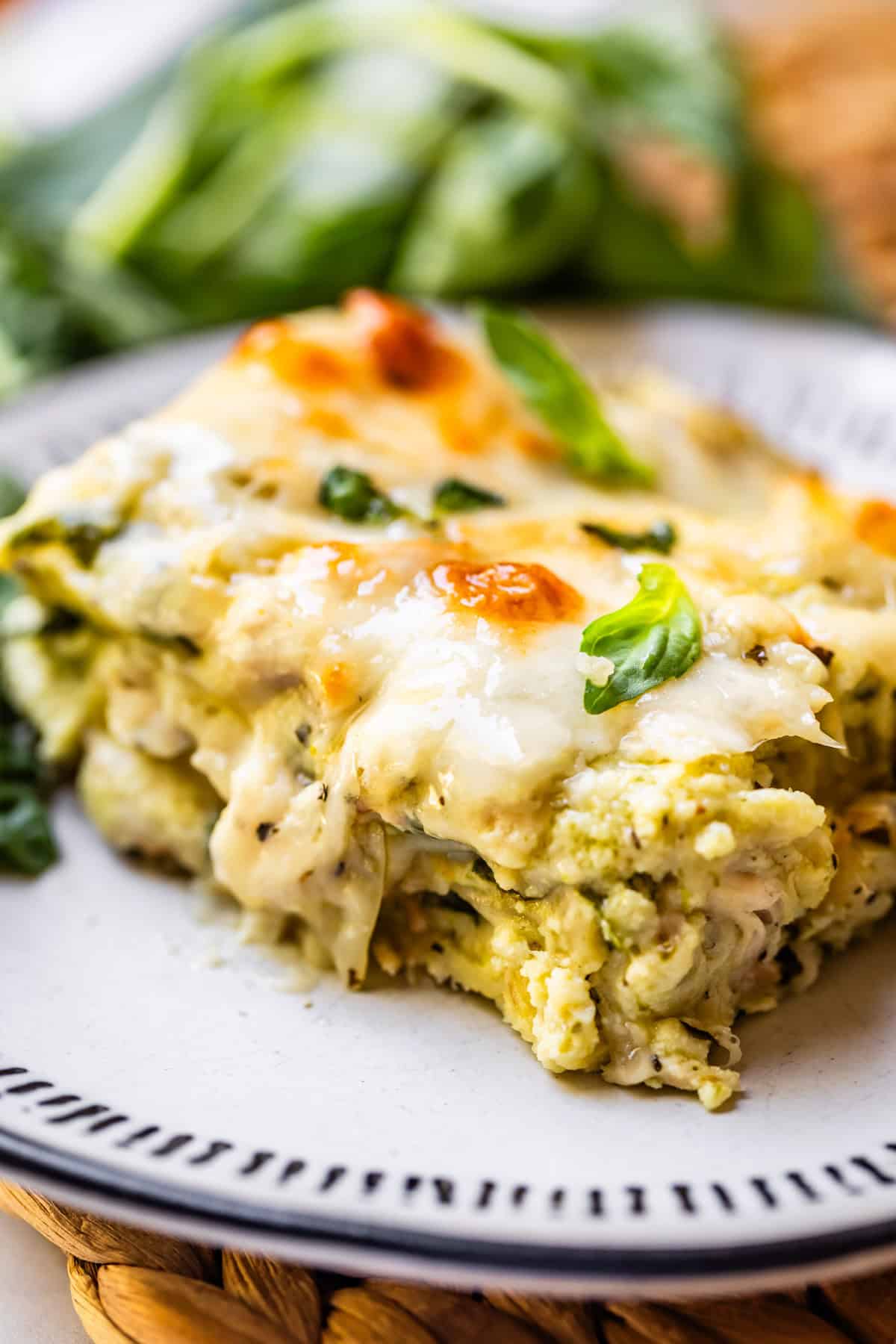 A close-up of a slice of creamy, cheesy vegetable lasagna garnished with fresh basil, served on a white plate with a decorative rim. Green leafy vegetables are blurred in the background.