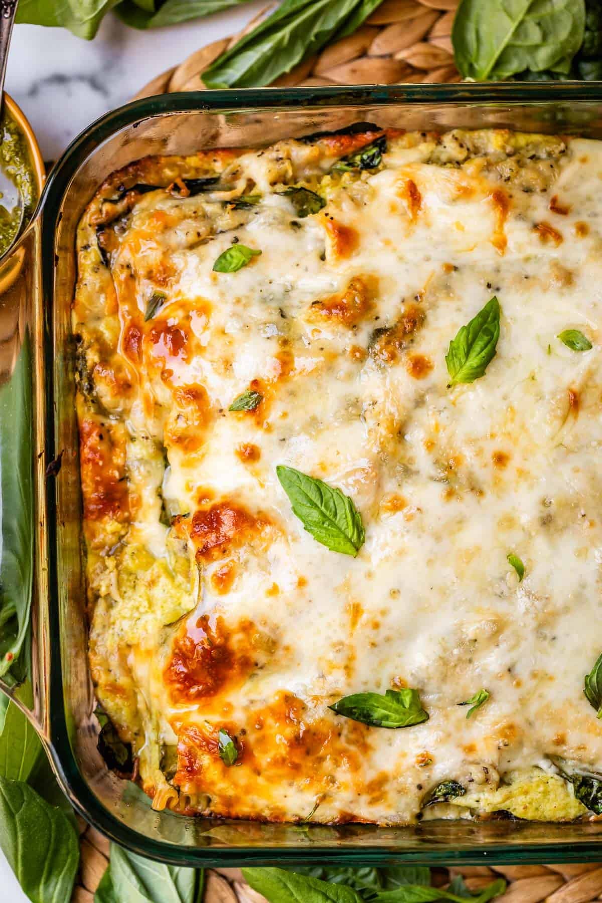 A close-up of a baked cheesy casserole in a glass dish, topped with melted golden cheese and fresh basil leaves, surrounded by fresh spinach leaves.