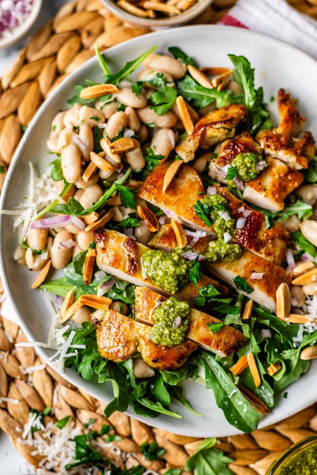 A plate with grilled chicken slices on a bed of arugula and white beans, topped with green pesto, toasted slivered almonds, grated cheese, and red onion slices.