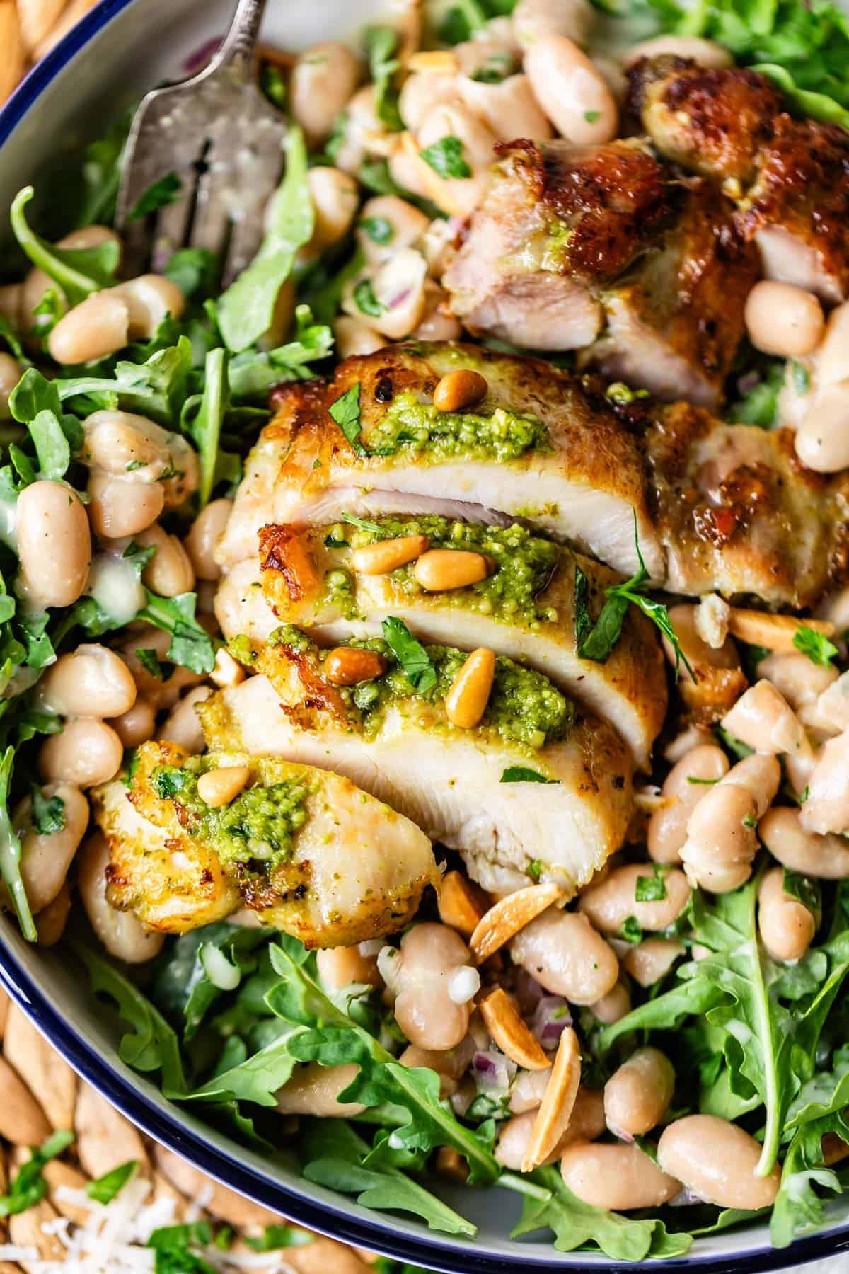 A plate of sliced grilled chicken topped with green pesto, served on a bed of arugula, white beans, pine nuts, and herbs, with a fork visible in the background.