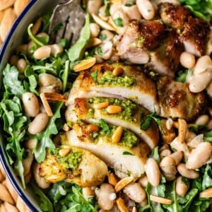A bowl filled with sliced grilled chicken topped with pesto, served on a bed of arugula, white beans, toasted pine nuts, and slivered almonds, with a fork resting on the side.