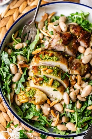 A bowl filled with sliced grilled chicken topped with pesto, served on a bed of arugula, white beans, toasted pine nuts, and slivered almonds, with a fork resting on the side.