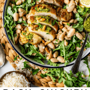 A bowl of arugula salad topped with sliced basil chicken, white beans, pine nuts, and dollops of pesto, garnished with lemon slices. Spoons and small bowls of cheese and pesto are nearby. Text reads: Basil Chicken Arugula Salad with White Beans.